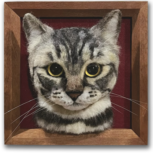 A realistic wool felt portrait of a tabby cat with large, expressive eyes, mounted in a wooden frame with a red background