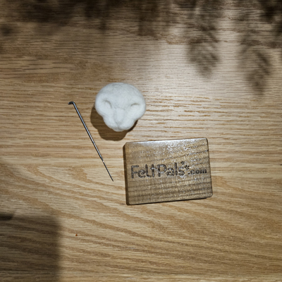 A felted wool base for a cat head alongside a felting needle and a wooden FeltPals.com logo block on a wooden surface.