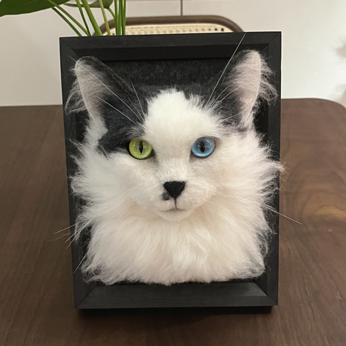 A realistic wool felt portrait of a long-haired cat with heterochromatic eyes (one green and one blue), mounted in a black frame.