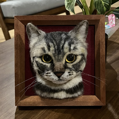 A realistic wool felt portrait of a tabby cat with large, expressive eyes and detailed facial markings, mounted in a wooden frame with a red background.