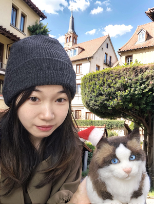 A woman wearing a gray beanie poses outdoors with a realistic wool felt sculpture of a Ragdoll cat, featuring blue eyes, in a picturesque setting with European-style buildings and manicured greenery.