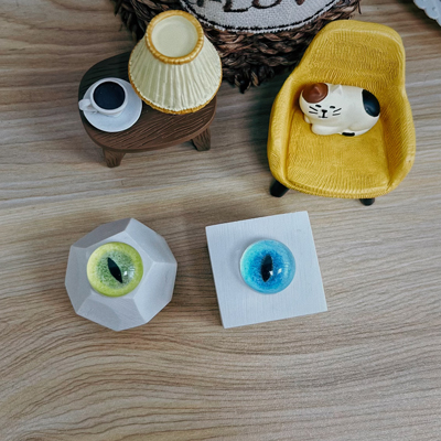 Two handmade cat eyes, one green and one blue, displayed on geometric stands on a wooden surface, with miniature furniture and a cat figurine in the background.