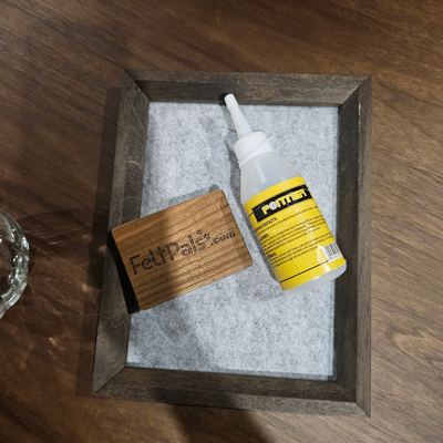 A wooden FeltPals.com logo block and a bottle of adhesive placed on a grey felt background within a dark wooden frame.