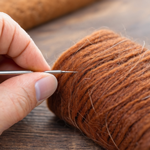 Close-up of a hand holding a needle, working on a roll of brown wool felt on a wooden surface.