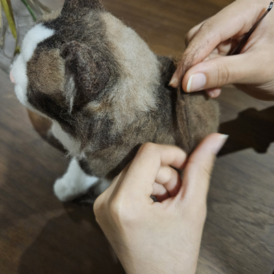 Hands working on a detailed wool felt sculpture of a cat, using a felting needle to shape and add texture to the fur.