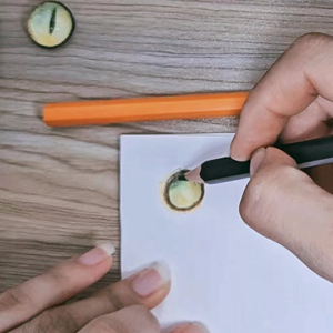 Close-up of a hand drawing a cat's eye with colored pencils on white paper, with a finished eye and an orange pencil nearby.