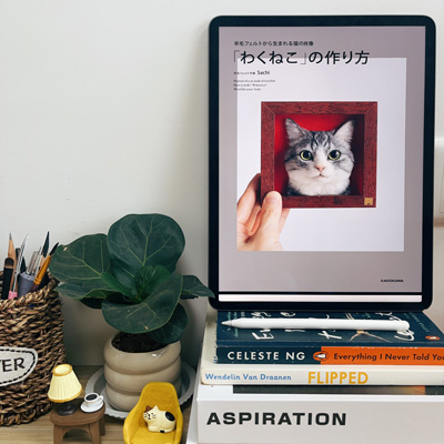 A workspace with a tablet displaying a Japanese needle-felting book cover, a stack of books, a plant, and a basket of pencils.