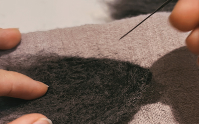 Close-up of hands working on needle felting a cat's ear using black wool on a grey fabric base.