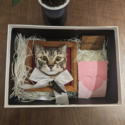 A packaged gift box containing a wool felt portrait of a tabby cat mounted in a wooden frame with a white bow, a FeltPals.com logo block, and a pink heart-shaped card.