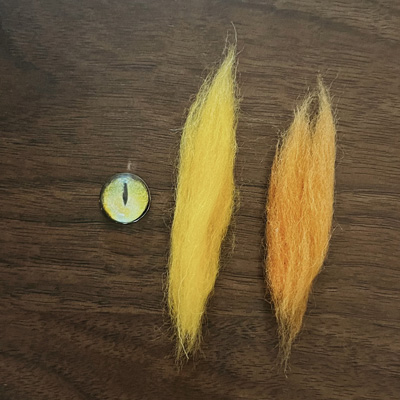 A yellow glass eye with a black vertical pupil and two tufts of wool, one yellow and one orange, placed on a wooden background.