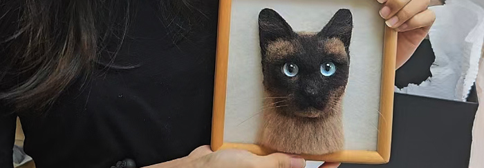 Needle-felted portrait of a Siamese cat in a wooden frame, held by a person.