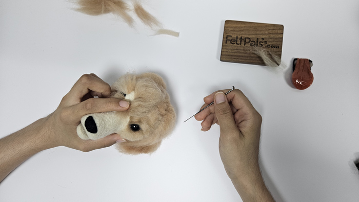 Hands crafting a felt dog head with a needle, surrounded by crafting tools on a white surface.