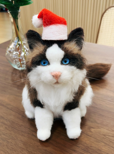 A wool felt sculpture of a Ragdoll cat with blue eyes wearing a red and white Santa hat, displayed on a wooden table with a vase in the background.