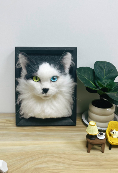 A wool felt portrait of a long-haired cat with heterochromatic eyes (one green and one blue) mounted in a black frame, displayed on a wooden surface next to a potted plant and miniature furniture.