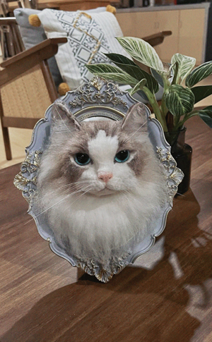 A realistic wool felt portrait of a Ragdoll cat with blue eyes, displayed in an ornate, vintage-style frame on a wooden table with a potted plant in the background.