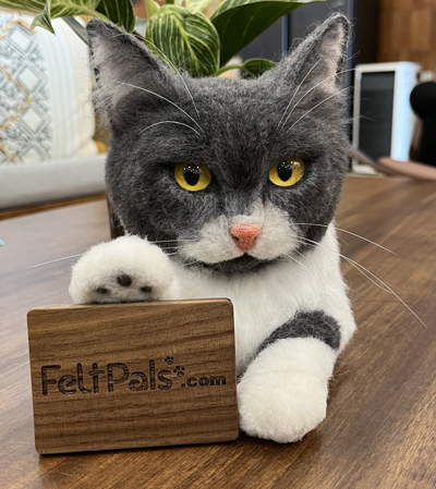 A realistic wool felt sculpture of a British Shorthair cat with yellow eyes, posed with its paw on a wooden FeltPals.com logo block, displayed on a wooden table.