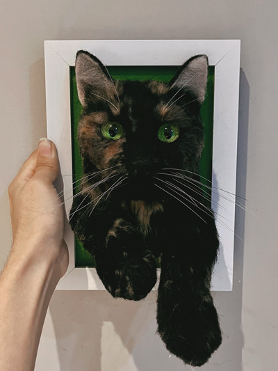 A realistic wool felt sculpture of a black cat with green eyes, mounted in a white frame with a green background, held by a hand.