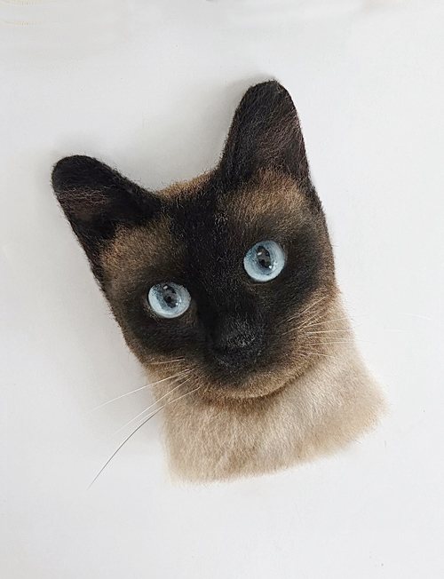 a highly realistic felt wool artwork of a Siamese cat, featuring blue eyes, dark facial markings, and light-colored fur.